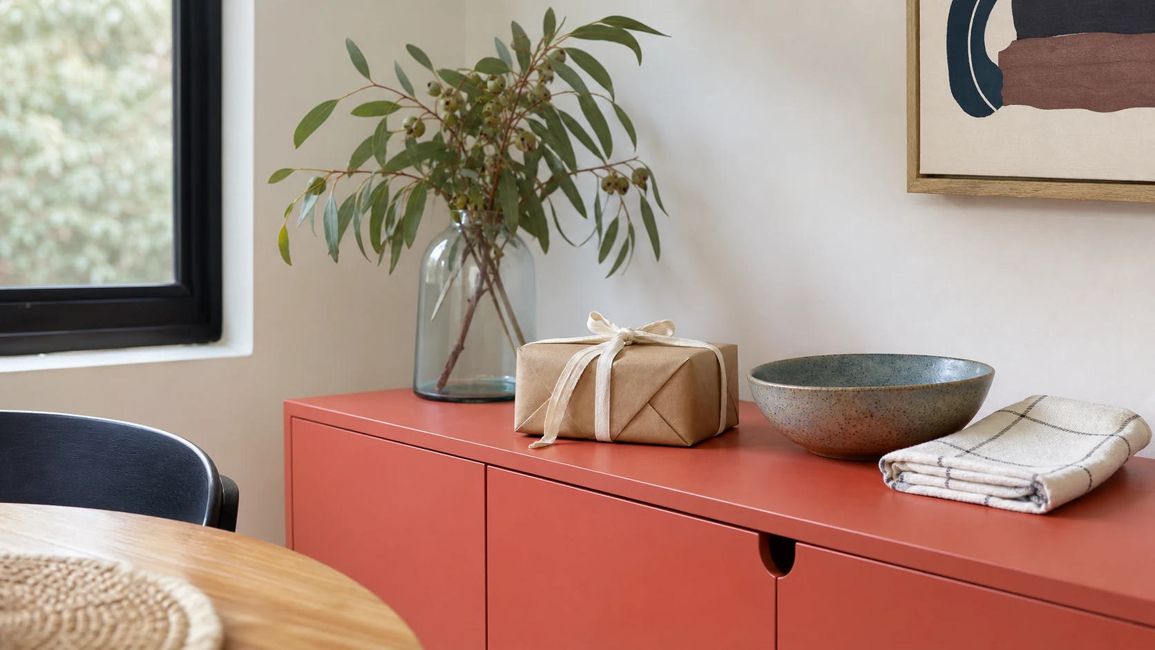 A modest home gift wrapped in kraft paper beside an unbranded ceramic bowl and folded cotton tea towel on a coral sideboard