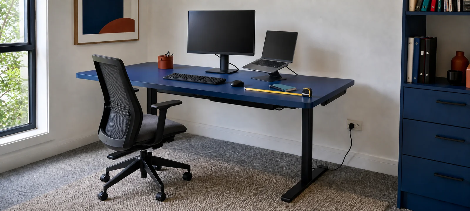 A cobalt blue home office desk with a monitor, laptop, chair clearance, measuring tape and nearby power point