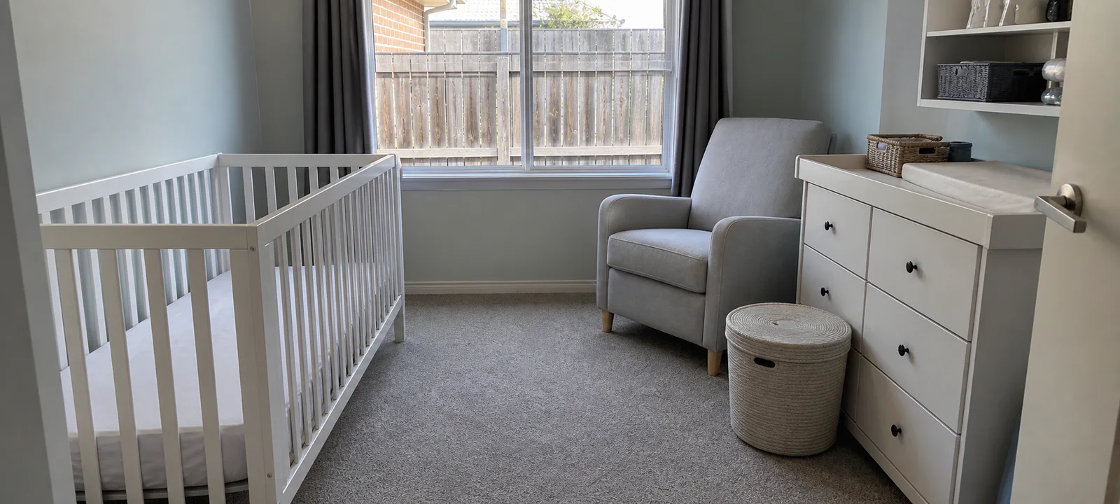 A functional nursery with an empty cot, change area, armchair, closed storage and a small laundry basket